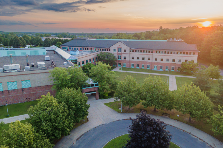 Aerial view of the Center for Advanced Material Processing (CAMP) at Clarkson University during sunset, with the building surrounded by trees and a circular driveway in the foreground. The warm colors of the sunset highlight the campus landscape, emphasizing the scenic environment.