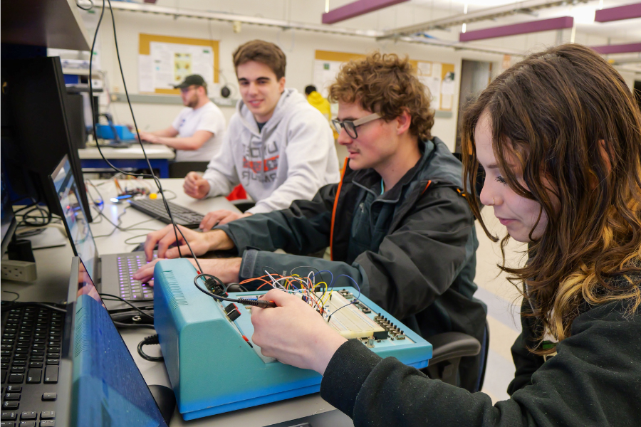 Clarkson University students working collaboratively in an electrical engineering lab, using equipment and laptops to build and test circuits. The environment showcases hands-on learning and teamwork in a technical academic setting.